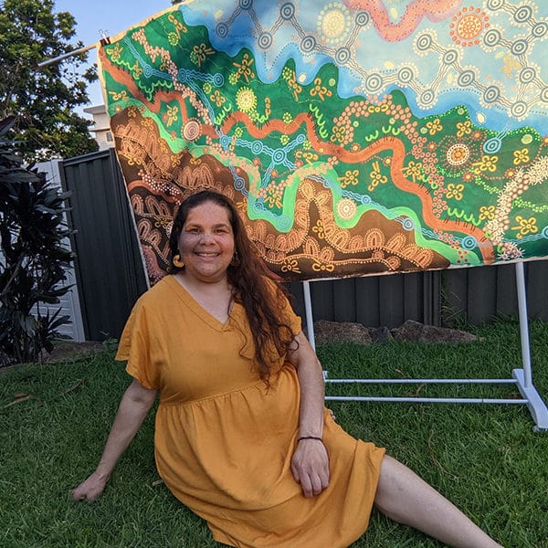 Aboriginal artist Leah Cummins sitting on the grass, smiling in front of one of her own large colourful artworks